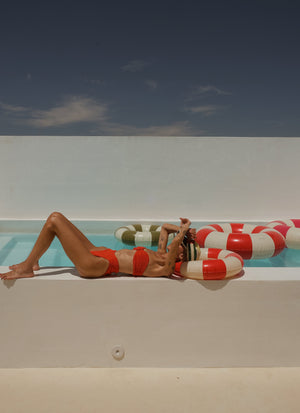 A woman in a red and white striped bikini lounges on the edge of a swimming pool under a clear blue sky, with a red and white striped pool float beside her.