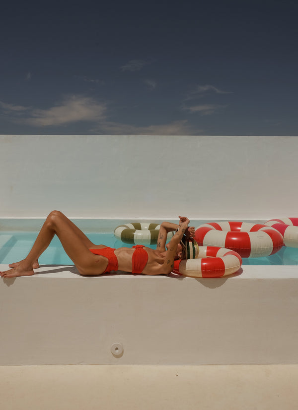 A woman in a red and white striped bikini lounges on the edge of a swimming pool under a clear blue sky, with a red and white striped pool float beside her.